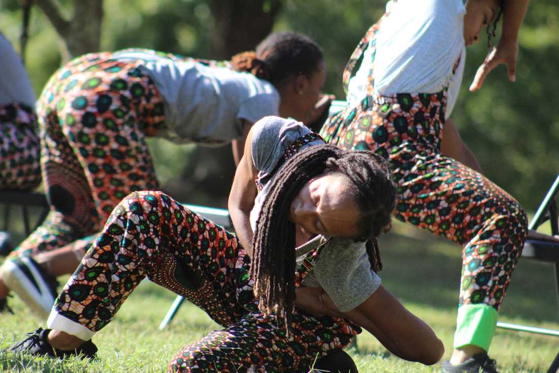 Four Black women dance in a field around black folding chairs. They are wearing orange, green, and black African wax print pants with suspenders and gray tshirst.