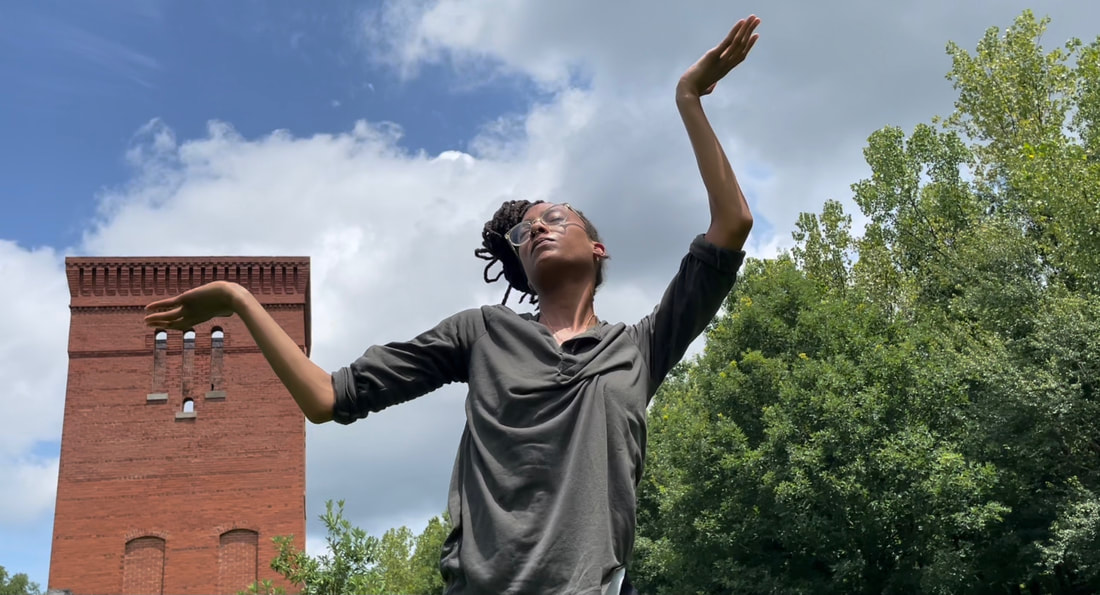 A Black women dances outside in front of a brick tower and green trees, eyes closed with her angular bent arms raised to the sky with open palms.