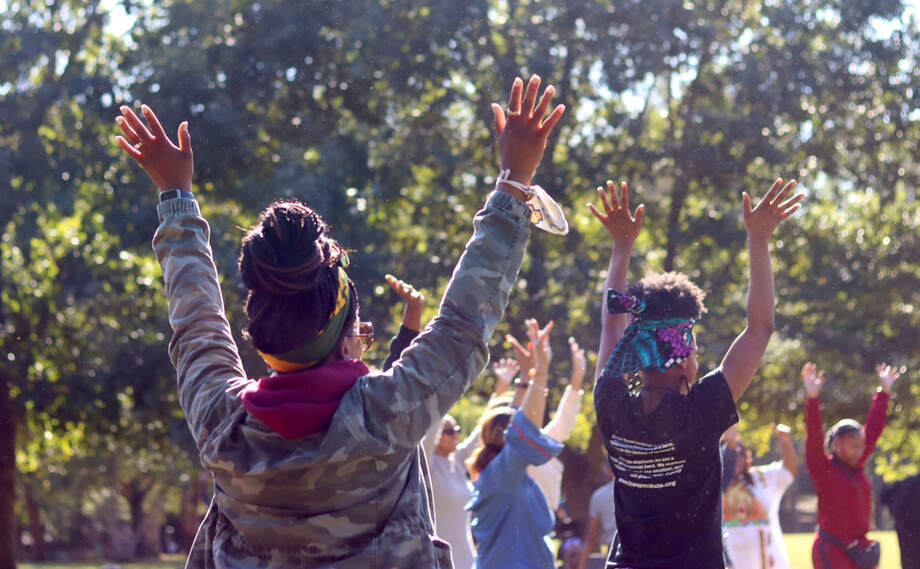 A group of people stand outside in a field on a sunny day with their arms raised to the sky. 