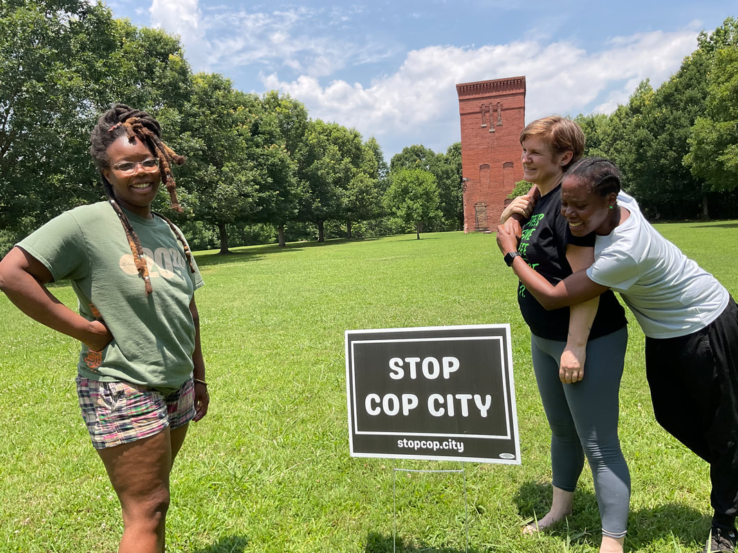 Three women stand in a green field next to a yard sign that reads “stop cop city.” They are smiling and laughing, a brick tower stands in the background.