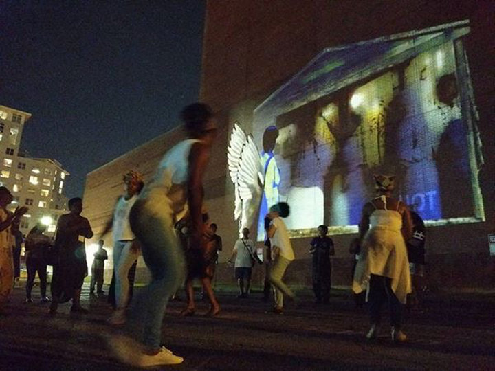 People gather, dance, and clap outside in a lot next to building at night with an image of an angel and ring shout dancers projected onto the wall.