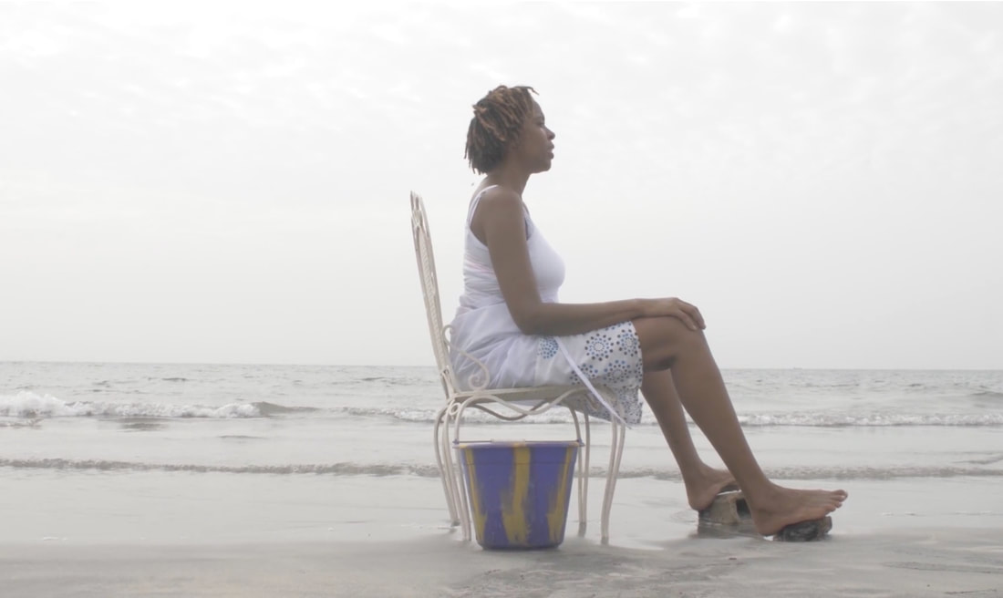 Charmaine sits in a chair on the beach. Waves in the background, blue bucket under the chair, her feet rest on rocks.