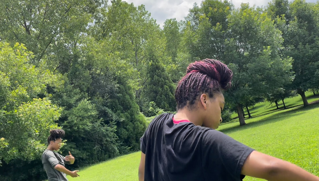 Grass field surrounded by green trees. Two Black women, with hair in locks twisted on top of their heads, dance in the middle of the field.